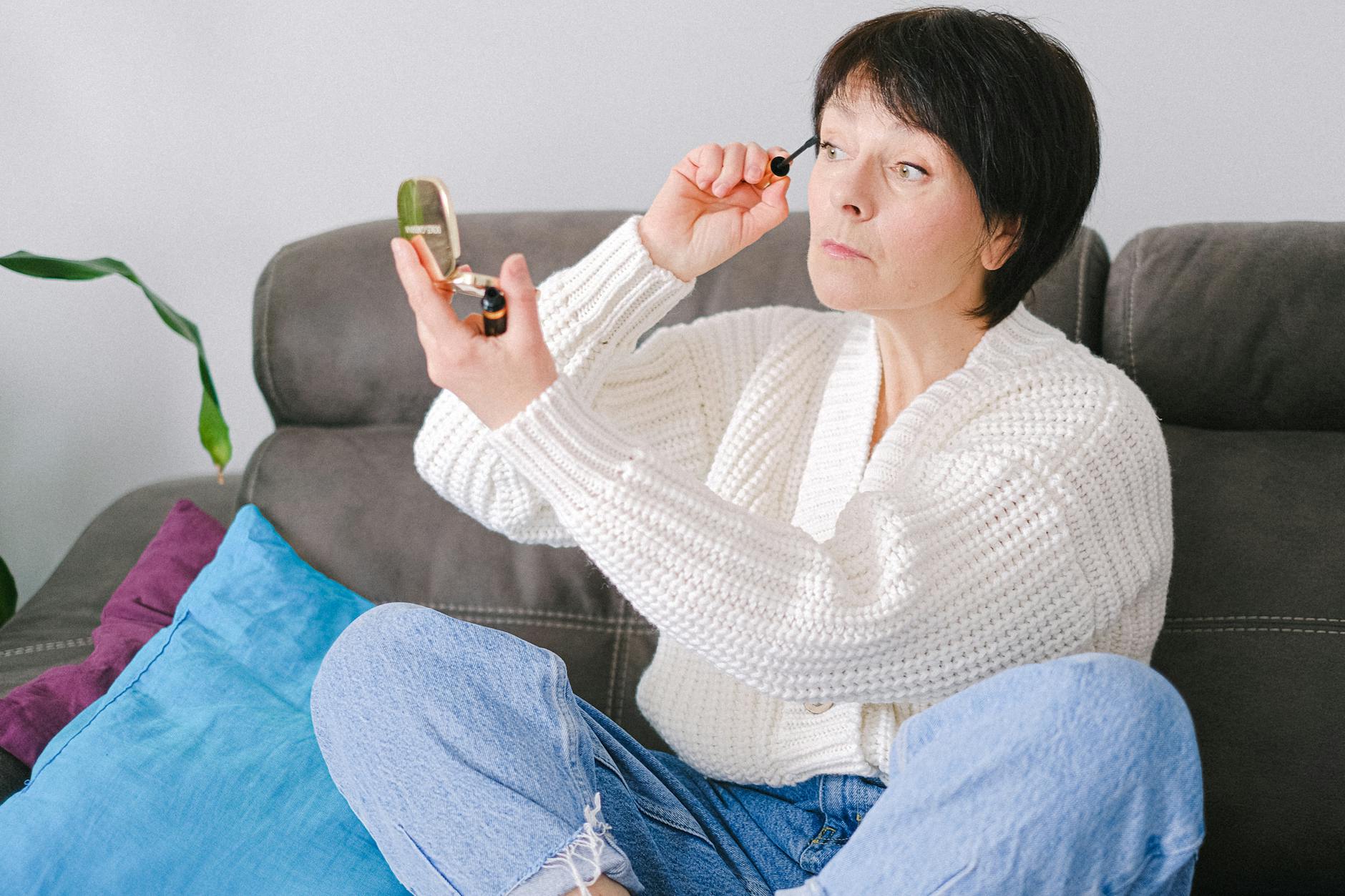 Starsza kobieta na kanapie maluje rzęsy tuszem w przytulnym wnętrzu